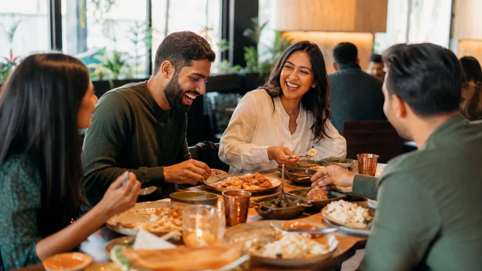 Friends enjoying a lively meal with South Asian dishes in a warmly lit restaurant dining room.