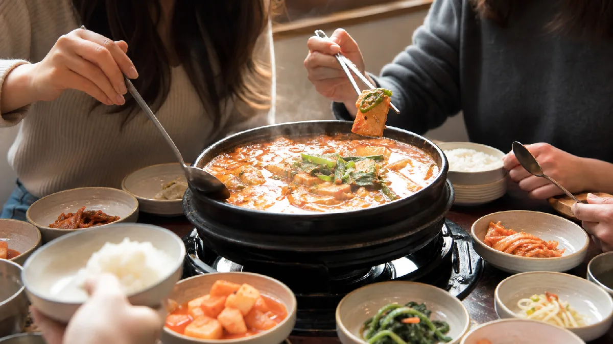 Friends gathered around a table sharing a bubbling Korean stew and assorted side dishes in a cozy dining room.