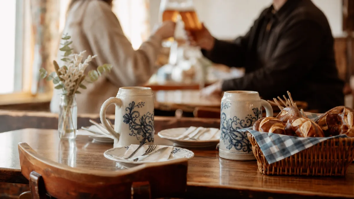 Cozy dining room with wooden table, traditional German beer steins, basket of pretzels, and couple toasting in the background.