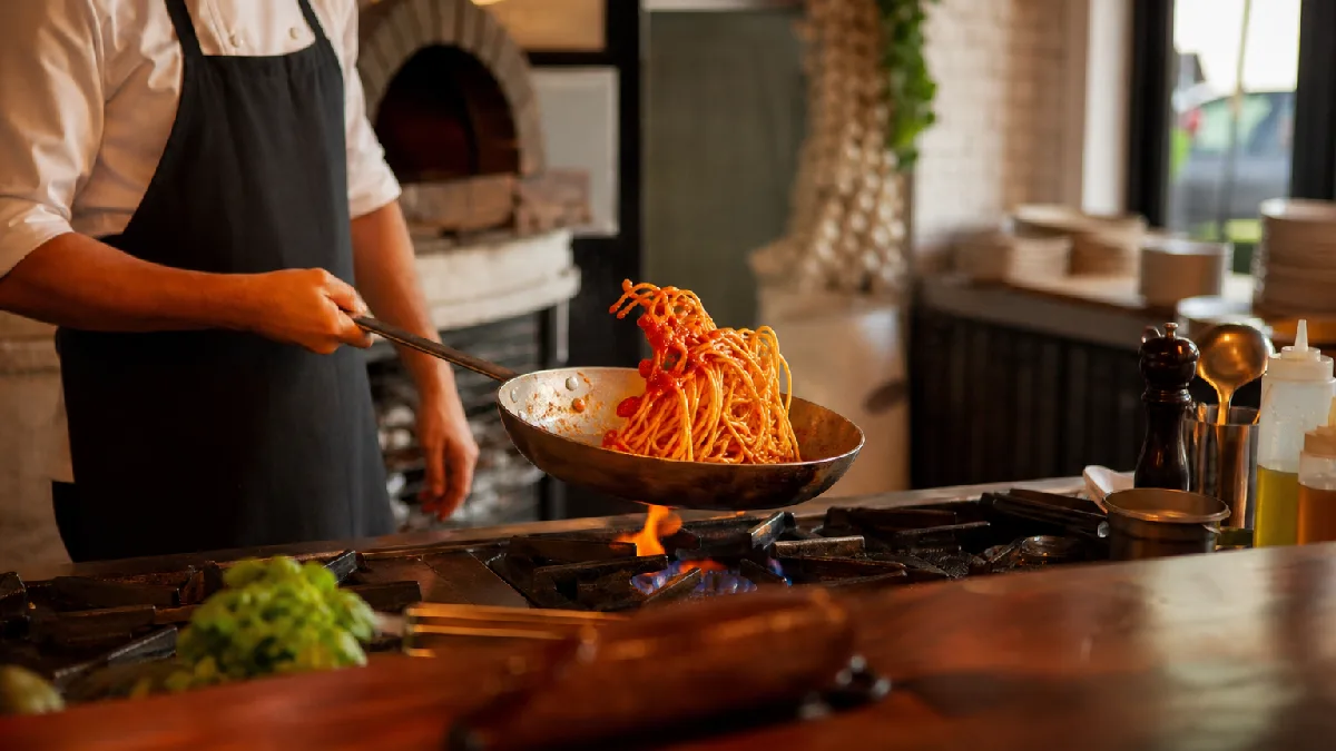 Cozy Italian-Mediterranean dining table with candles and wine near windows showing a chef cooking pasta in the background.