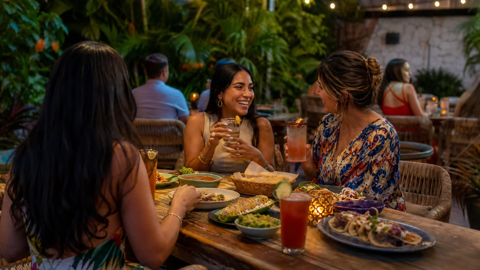 Friends dining outdoors at a cozy patio in warm evening light with colorful Mexican and Latin American dishes on the table.